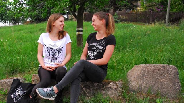Zwei Schülerinnen des Start-up-Unternehmens "Diverse" sitzen auf Steinen im Grünen und tragen die selbst entworfenen T-Shirts