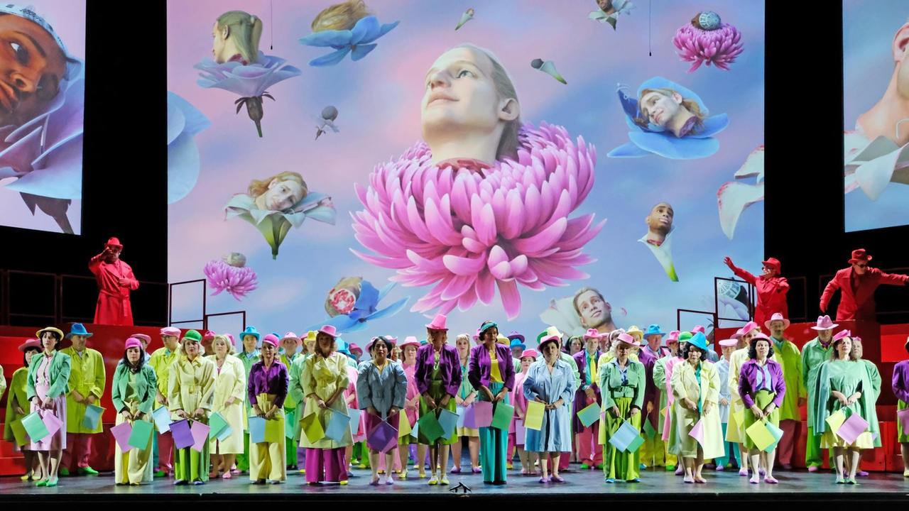 Szenenfoto aus Turandot: Der knallbunt gekleidete Staatsopernchor vor einer riesigen Projektion. Darauf zu sehen sind in Blüten platzierte Männerköpfe. Szenenfoto aus Turandot: Der knallbunt gekleidete Staatsopernchor vor einer riesigen Projektion. Darauf zu sehen sind in Blüten platzierte Männerköpfe.