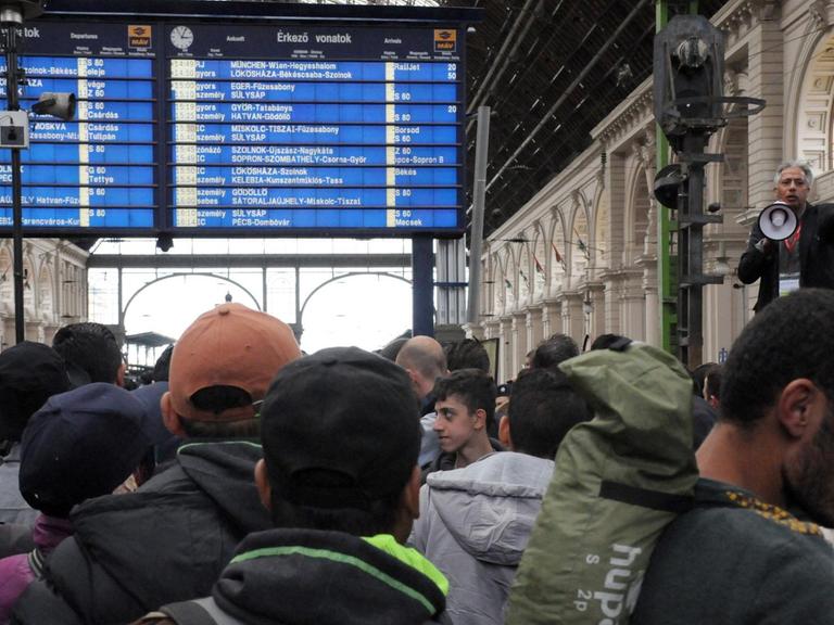 Flüchtlinge stehen auf dem Ostbahnhof in Budapest und hören einem Man mit Megafon zu.
