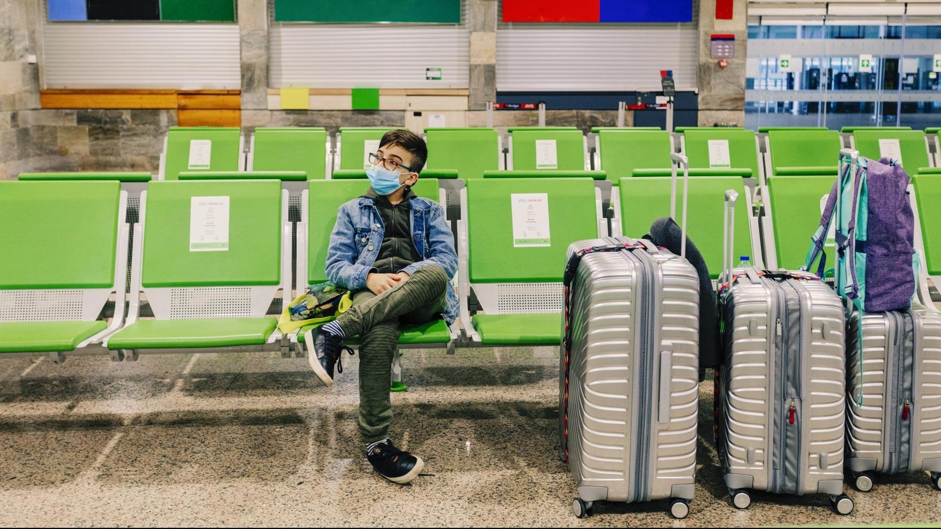 Ein junge sitzt mit Maske in einem Flugzeug-Terminal mit großen Gepäck (Symbolfoto)
