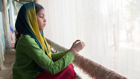 Das Bild zeigt am linken Bildrand ein Mädchen aus Indien, in grünem Gewand, bei ihrer täglichen Arbeit am Webrahmen. Dieses Foto ist das Motiv des aktuellen Plakats der Aktion Dreikönigssingen 2018.