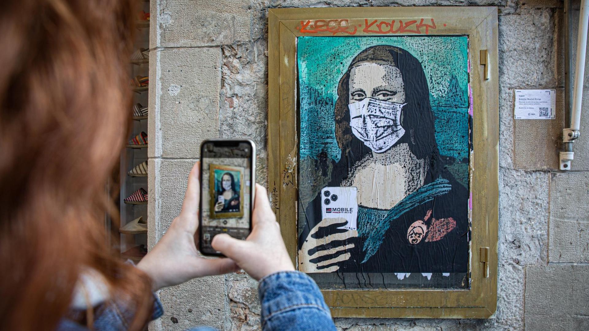 An einer Wand ist eine Graffito-Adaption der Mona Lisa zu sehen. Sie trägt Mundschutz und hält ein Smartphone, als ob sie fotografieren würde. Im Vordergrund sind die Hände einer Frau zu sehen, die dieses Bild fotografieren.