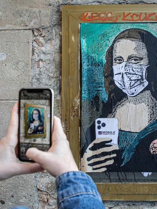 An einer Wand ist eine Graffito-Adaption der Mona Lisa zu sehen. Sie trägt Mundschutz und hält ein Smartphone, als ob sie fotografieren würde. Im Vordergrund sind die Hände einer Frau zu sehen, die dieses Bild fotografieren.