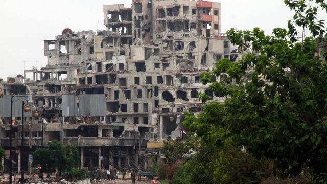 Ein durch Bomben zerstörter Wohnblock in Homs, Syrien im Mai 2014. Soldaten stehen vor der Ruine. Bäume säumen die Straße