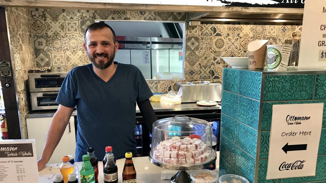 Ein lächelnder Mann in T-Shirt und mit dunklem Vollbart steht hinter einem Tresen auf dem Plastikflaschen und Schachteln stehen. Ein lächelnder Mann in T-Shirt und mit dunklem Vollbart steht hinter einem Tresen auf dem Plastikflaschen und Schachteln stehen.