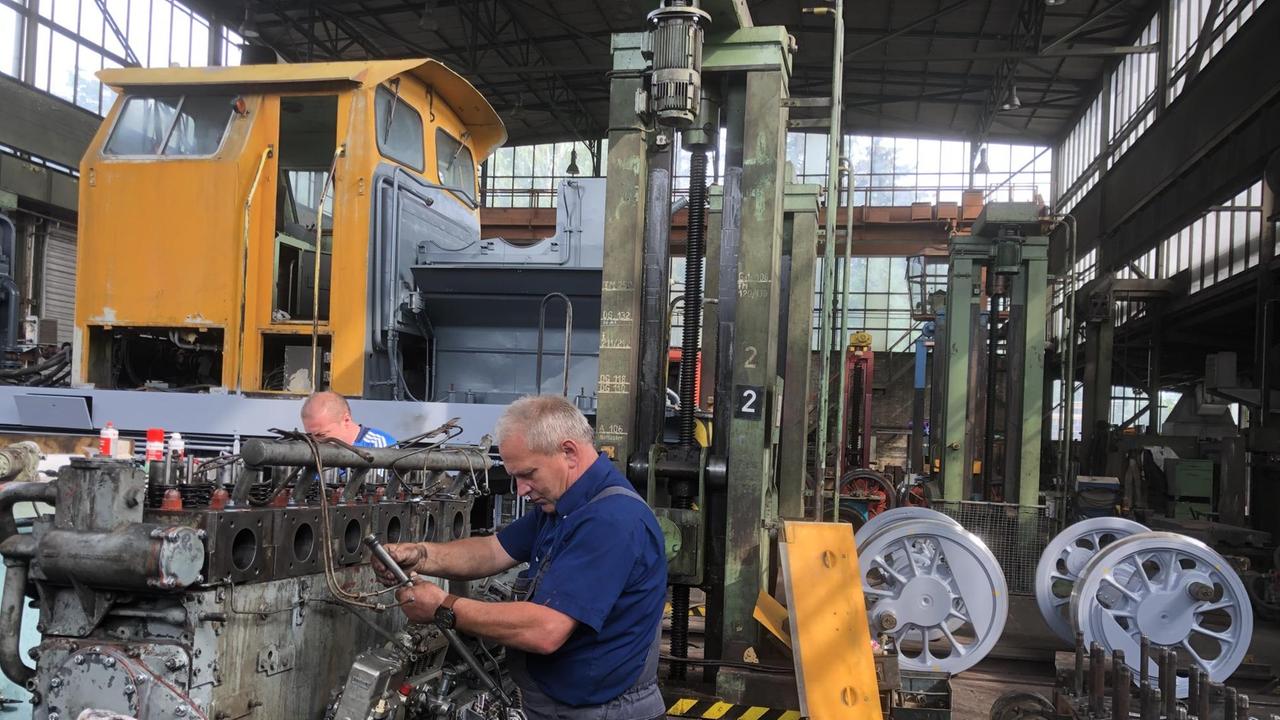In einer Fabrikhalle steht ein Arbeiter an einer Lokomotive mit einem orangen Fahrerhaus.