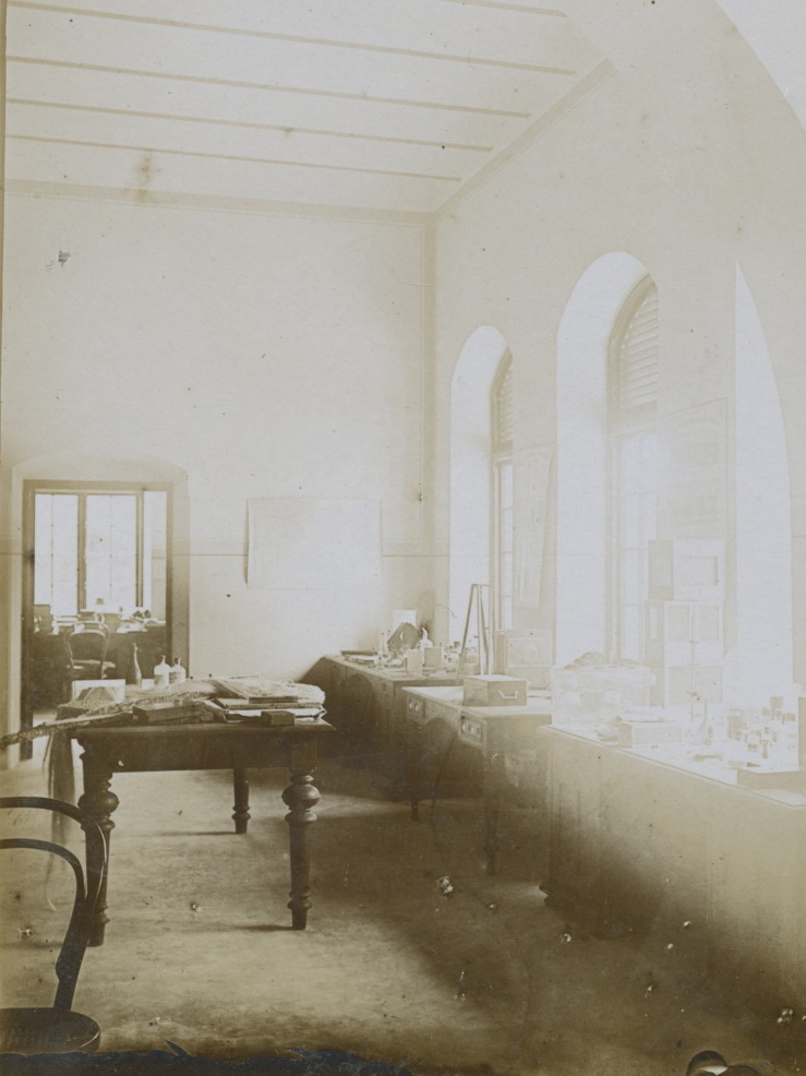 Ein hoher Raum mit weißen Wänden, ein Tisch vor Fenstern mit Rundbögen, das Arbeitszimmer des Kolonialbeamten Franz Stuhlmann in Deutsch-Ostafrika im Jahr 1900.