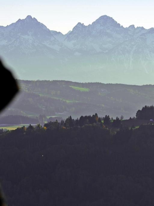 Ein Mann, der sich mit der rechten Hand vor dem Sonnenlicht schützt, schaut von Hohenpeißenberg aus auf die Alpen.