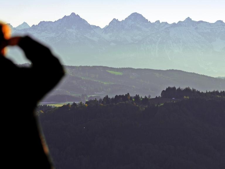 Ein Mann, der sich mit der rechten Hand vor dem Sonnenlicht schützt, schaut von Hohenpeißenberg aus auf die Alpen.