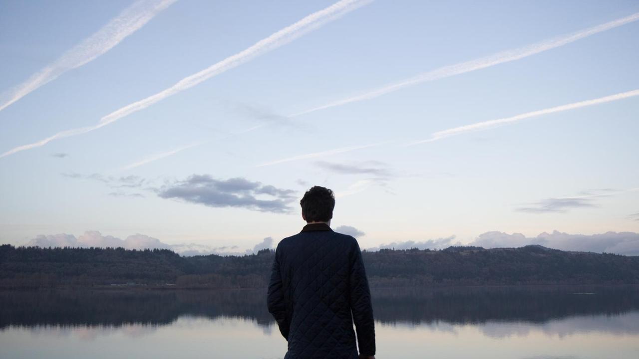 Ein Mann steht mit dem Rücken zum Betrachter vor einem See. Am Himmel sind Kondenstreifen zu sehen. Ein Mann steht mit dem Rücken zum Betrachter vor einem See. Am Himmel sind Kondenstreifen zu sehen.