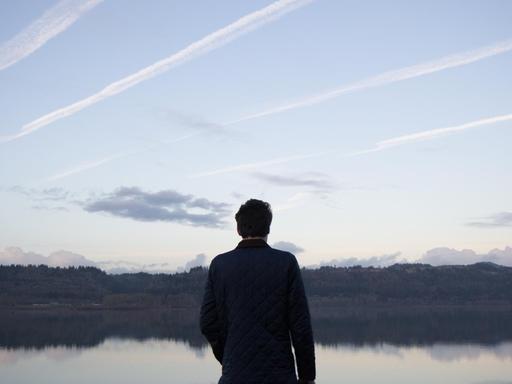 Ein Mann steht mit dem Rücken zum Betrachter vor einem See. Am Himmel sind Kondenstreifen zu sehen.