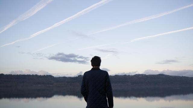 Ein Mann steht mit dem Rücken zum Betrachter vor einem See. Am Himmel sind Kondenstreifen zu sehen.