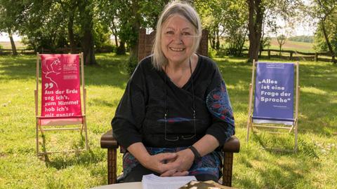 Helga Schubert sitzt auf einer Wiese in einem Stuhl, im Hintergrund sind Bäume zu sehen.