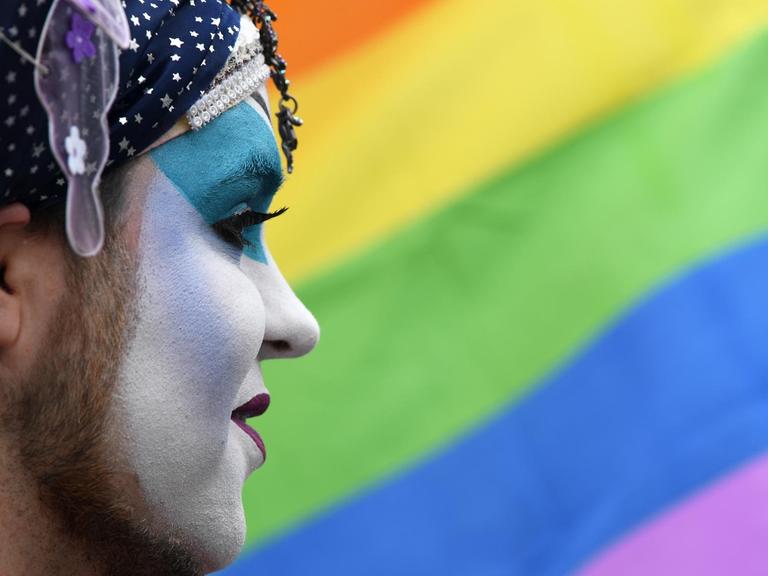 Bärtiger geschminkter Mann im Profil vor einer Regenbogenflagge.