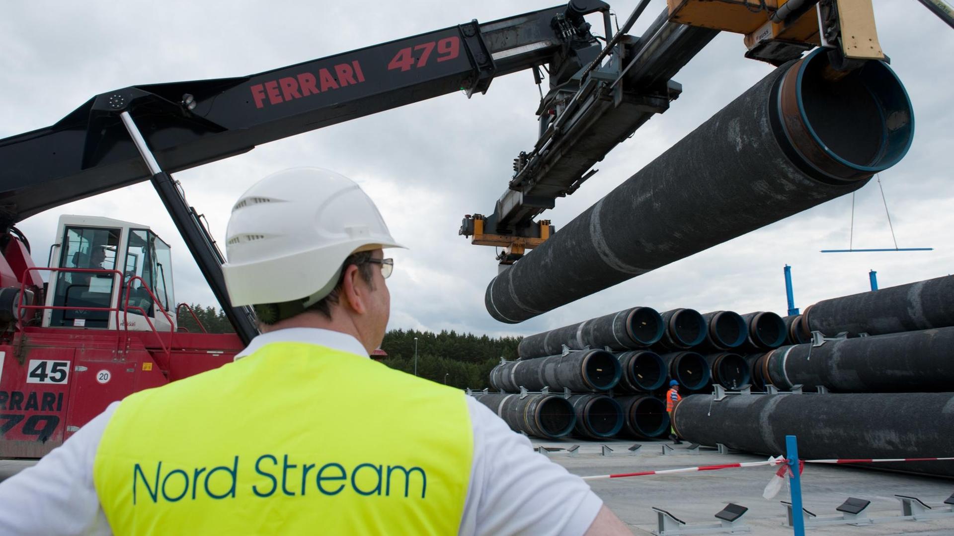 Im Vordergrund steht ein Mann in gelber Warnweste und beobachtet einen Bagger, der Pipelinerohre hebt. Im Vordergrund steht ein Mann in gelber Warnweste und beobachtet einen Bagger, der Pipelinerohre hebt.