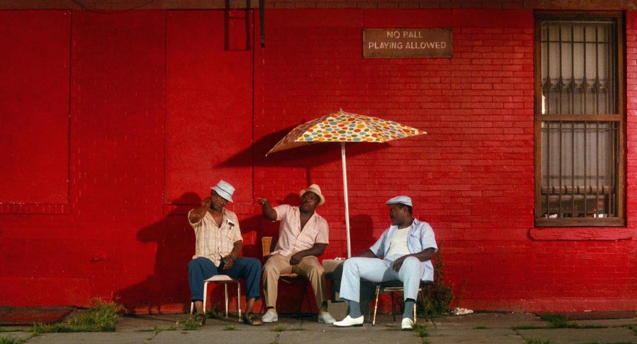 Szene aus dem Film "Do The Right Thing" von Regisseur Spike Lee aus dem Jahr 1989. Vor einer roten Hauswand sitzen bei glühender Hitze drei Männer unter einem Sonnenschirm. Szene aus dem Film "Do The Right Thing" von Regisseur Spike Lee aus dem Jahr 1989. Vor einer roten Hauswand sitzen bei glühender Hitze drei Männer unter einem Sonnenschirm.