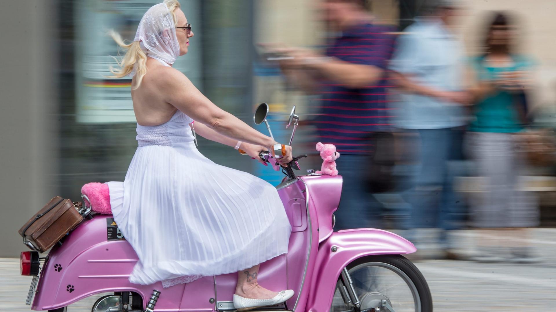 Ein Frau mit blonden Haaren im weißen Kleid und mit weißem Kopftuch fährt auf einer pinken "Schwalbe", dem Kult-Roller der DDR. Ein Frau mit blonden Haaren im weißen Kleid und mit weißem Kopftuch fährt auf einer pinken "Schwalbe", dem Kult-Roller der DDR.