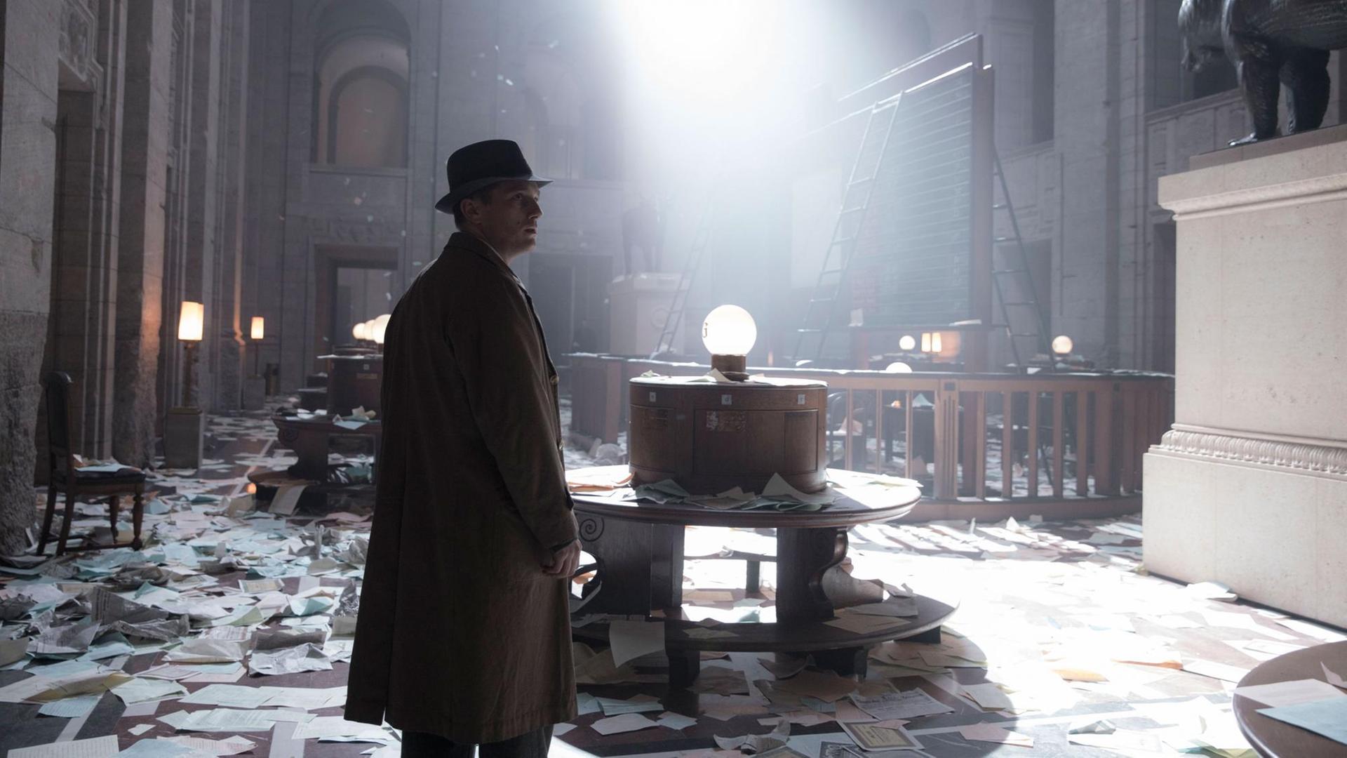 Ein Mann in einem Trench-Coat mit Hut steht in einem hohen Raum, der verwüstet wurde, überall liegen Blätter auf dem Boden. Ein Mann in einem Trench-Coat mit Hut steht in einem hohen Raum, der verwüstet wurde, überall liegen Blätter auf dem Boden.