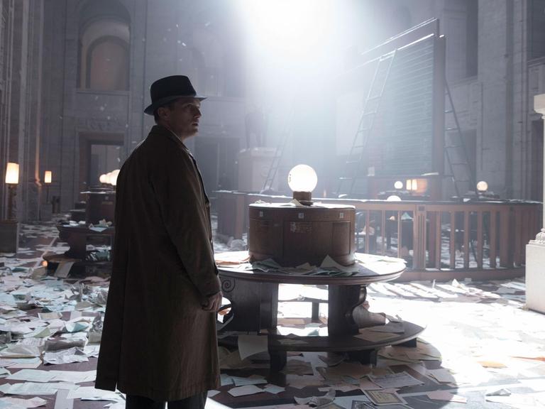 Ein Mann in einem Trench-Coat mit Hut steht in einem hohen Raum, der verwüstet wurde, überall liegen Blätter auf dem Boden.
