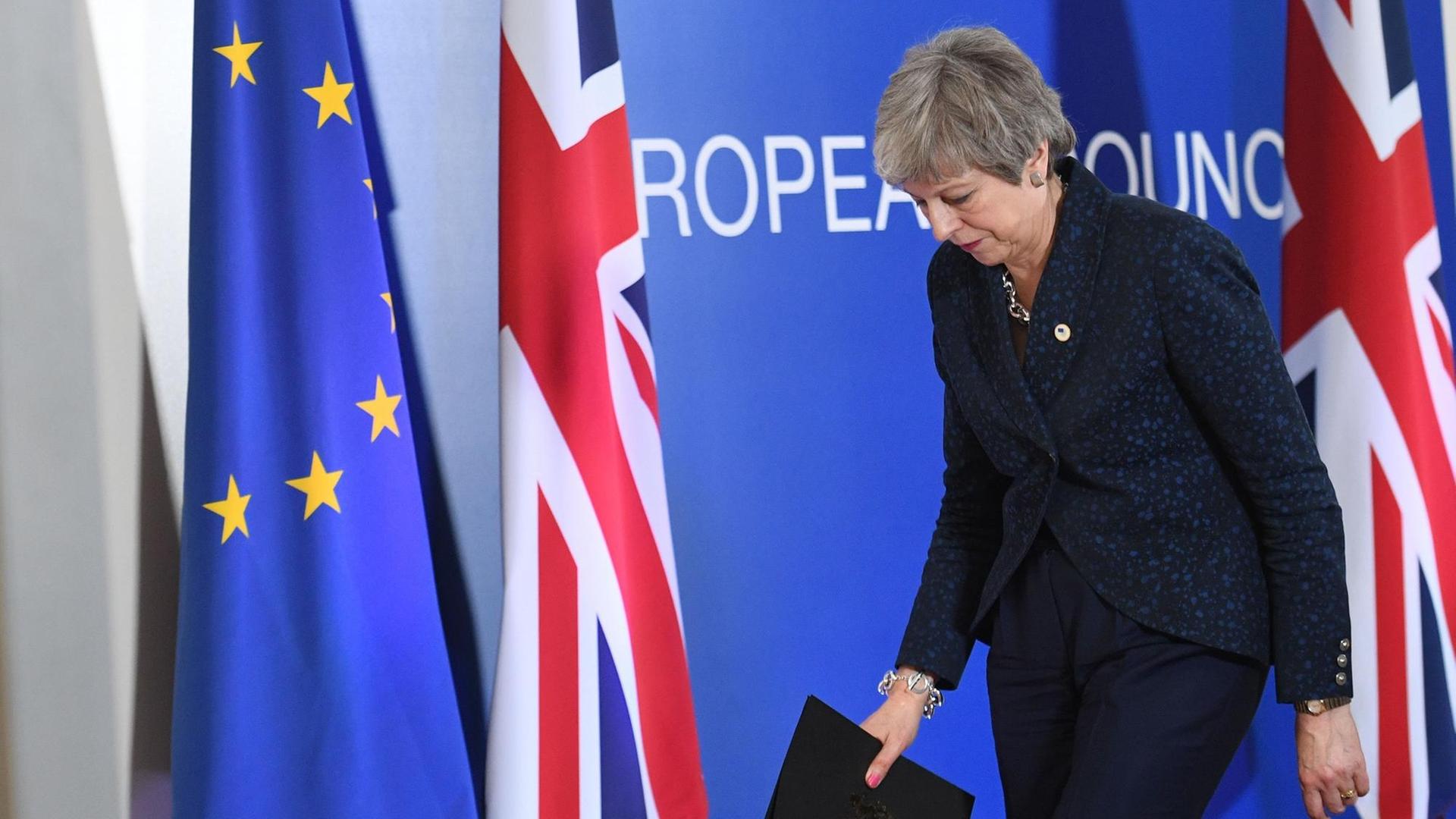 Theresa May verlässt den Raum, nachdem sie ein Statement abgegeben. Ihr Kopf ist gesenkt, im Hintergund sind die Flaggen der EU und Großbritanniens zu sehen. Theresa May verlässt den Raum, nachdem sie ein Statement abgegeben. Ihr Kopf ist gesenkt, im Hintergund sind die Flaggen der EU und Großbritanniens zu sehen.