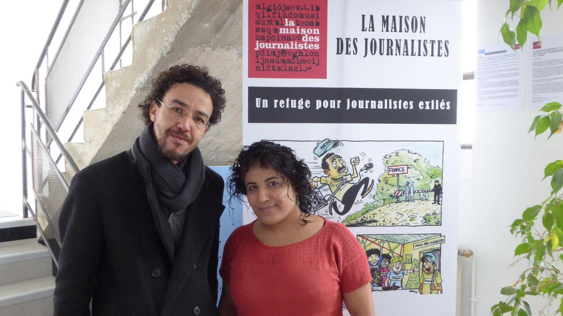 Ein Mann und eine Frau stehen in einem Treppenhaus. Hinter ihnen hängt ein Plakat mit der Aufschrift "La Maison des Journalistes" - "Das Haus der Journalisten"