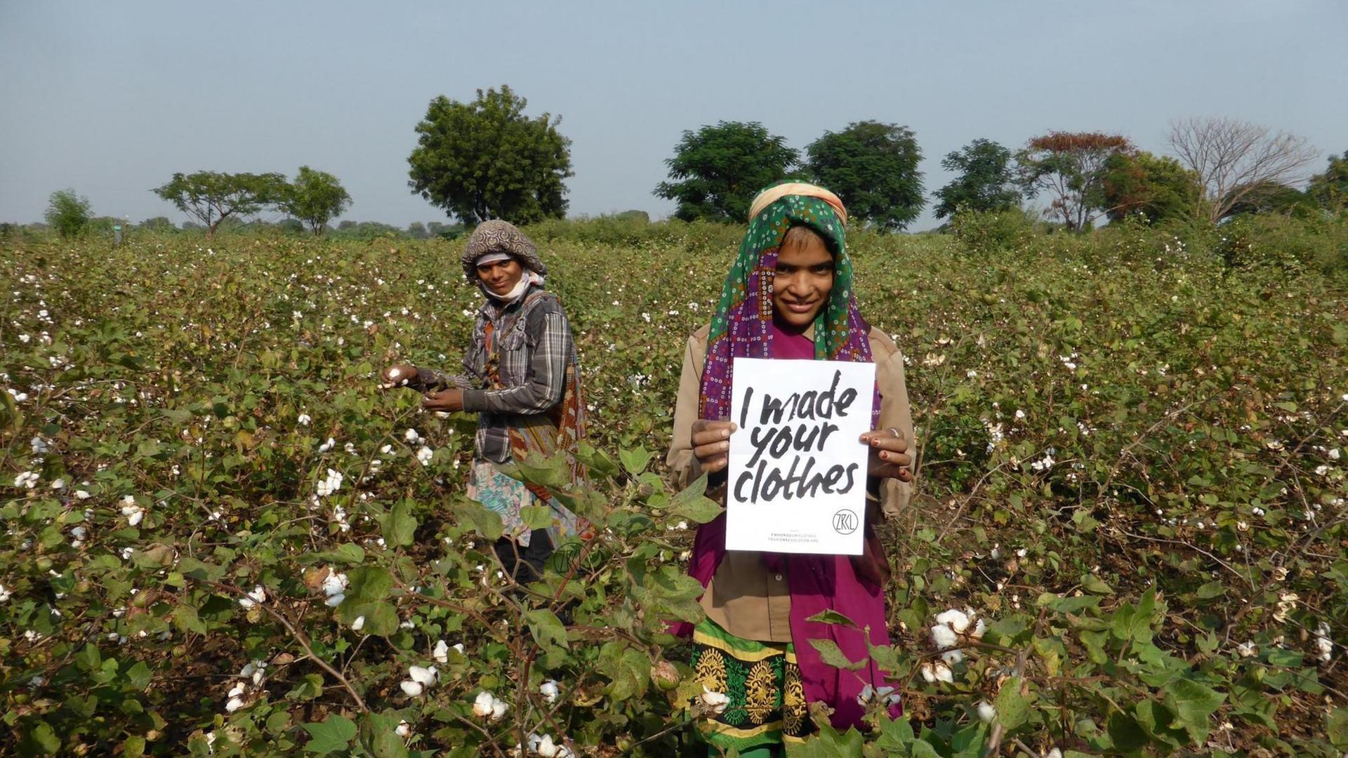 Zwei Arbeiterinnen auf einem Baumwollfeld halten ein Schild hoch auf dem steht "ich habe deine Kleidung gemacht".
