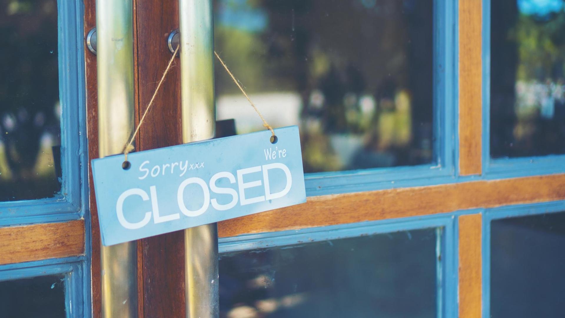 Ein Schild mit der Aufschrift "Sorry, we're closed" hängt an einer Ladentür. Ein Schild mit der Aufschrift "Sorry, we're closed" hängt an einer Ladentür.
