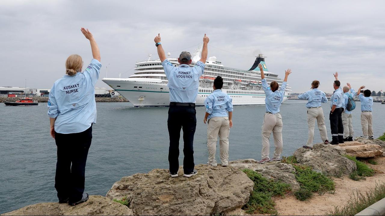 Menschen in Berufskleidung stehen mit Abstand zueinander am Ufer aufgereiht und winken dem abfahrenden Schiff Menschen in Berufskleidung stehen mit Abstand zueinander am Ufer aufgereiht und winken dem abfahrenden Schiff