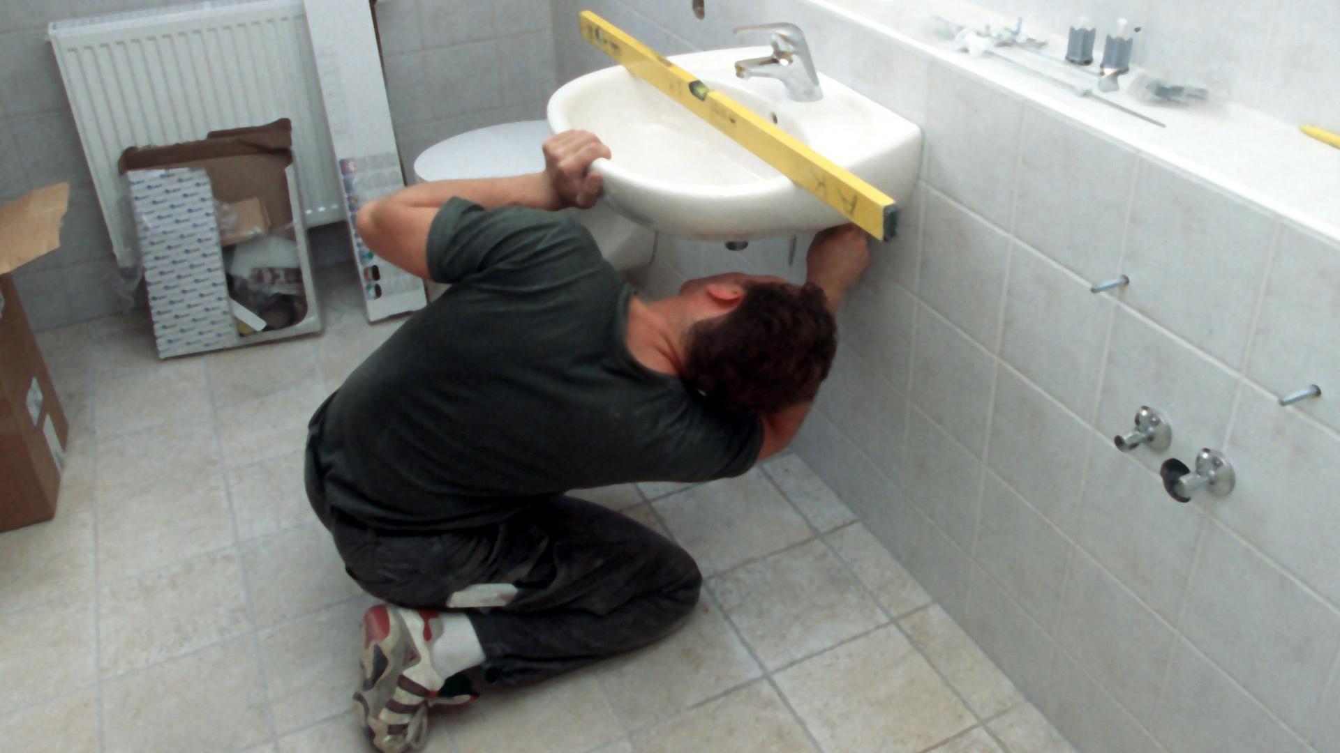 Ein Installateur kniet bei der Montage eines Waschbeckens im Badezimmer auf dem Boden. Mit der Wasserwaage tariert der Sanitärfachmann die korrekte Lage des Beckens aus. Ein Installateur kniet bei der Montage eines Waschbeckens im Badezimmer auf dem Boden. Mit der Wasserwaage tariert der Sanitärfachmann die korrekte Lage des Beckens aus.