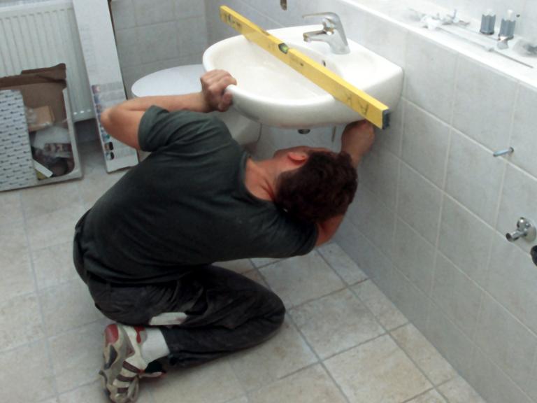 Ein Installateur kniet bei der Montage eines Waschbeckens im Badezimmer auf dem Boden. Mit der Wasserwaage tariert der Sanitärfachmann die korrekte Lage des Beckens aus. 