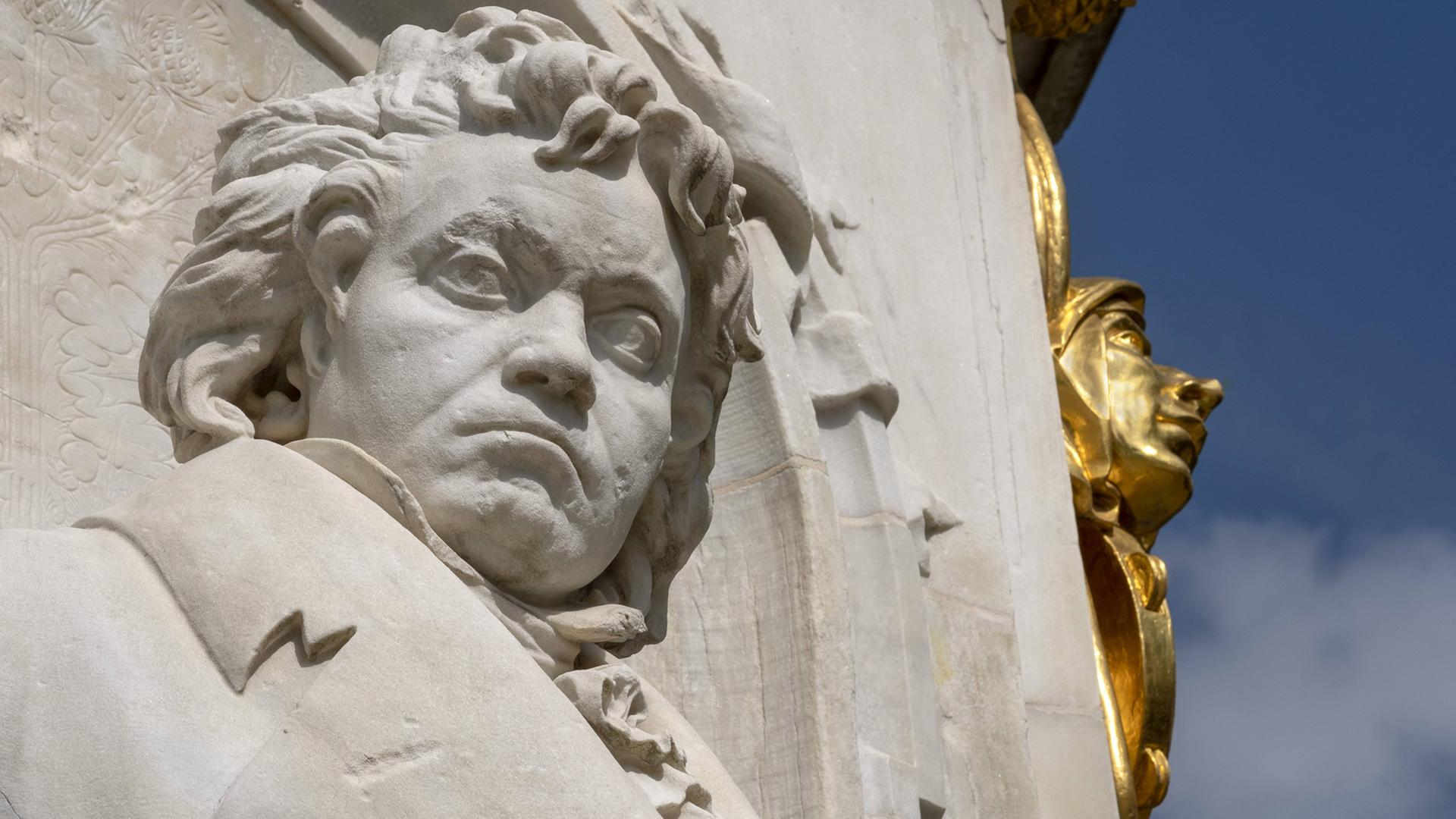Ein Denkmal von Ludwig van Beethoven aus hellem Stein, zu sehen ist er von Kopf bis Brust, im Hintergrund ein vergoldeter Teil des Denkmals und blauer Himmel.