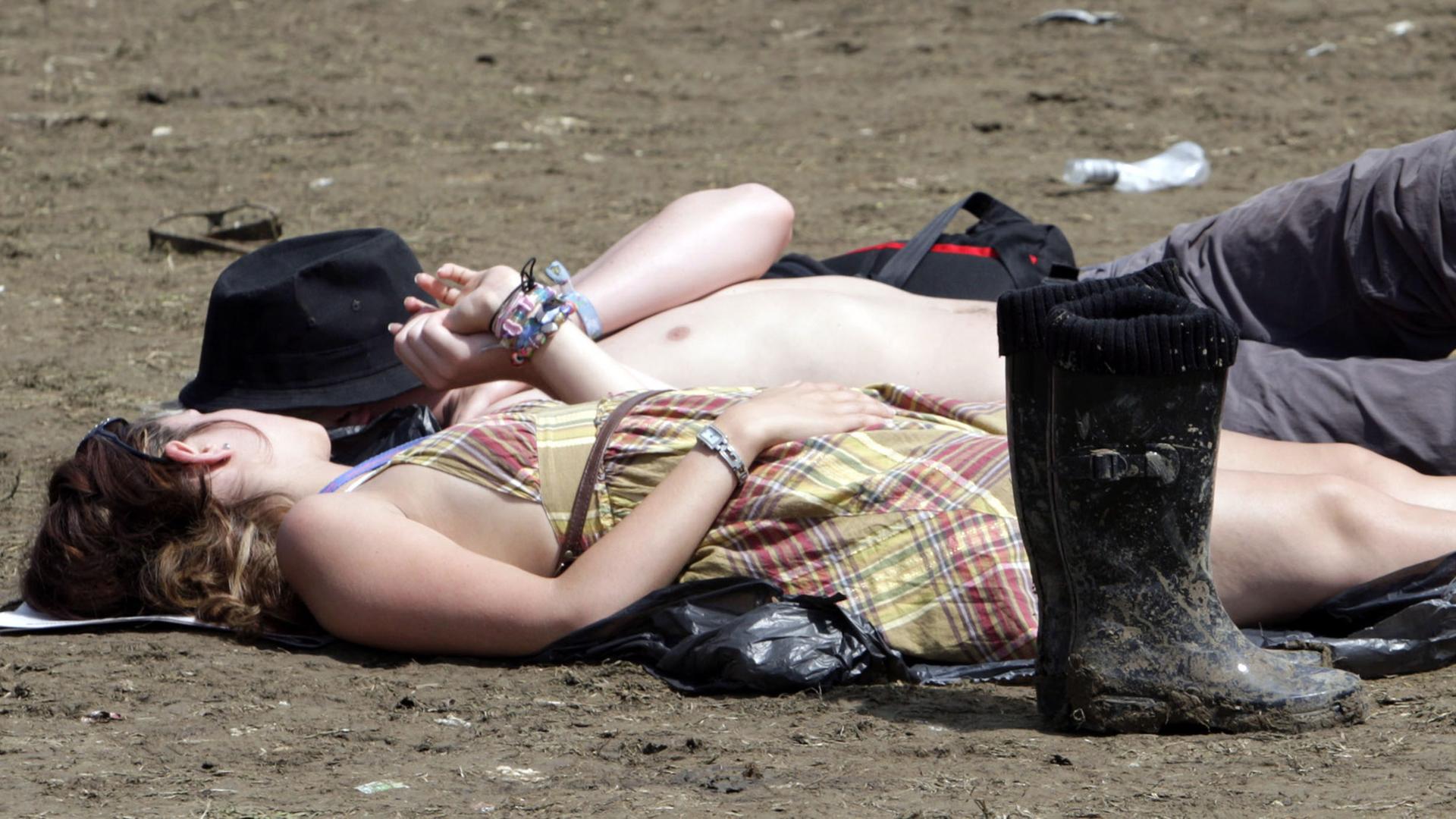 Ein Pärchen liegt auf dem erdigen Gelände des britischen Glastonbury-Festivals, im Vordergrund stehen Gummistiefel. Ein Pärchen liegt auf dem erdigen Gelände des britischen Glastonbury-Festivals, im Vordergrund stehen Gummistiefel.