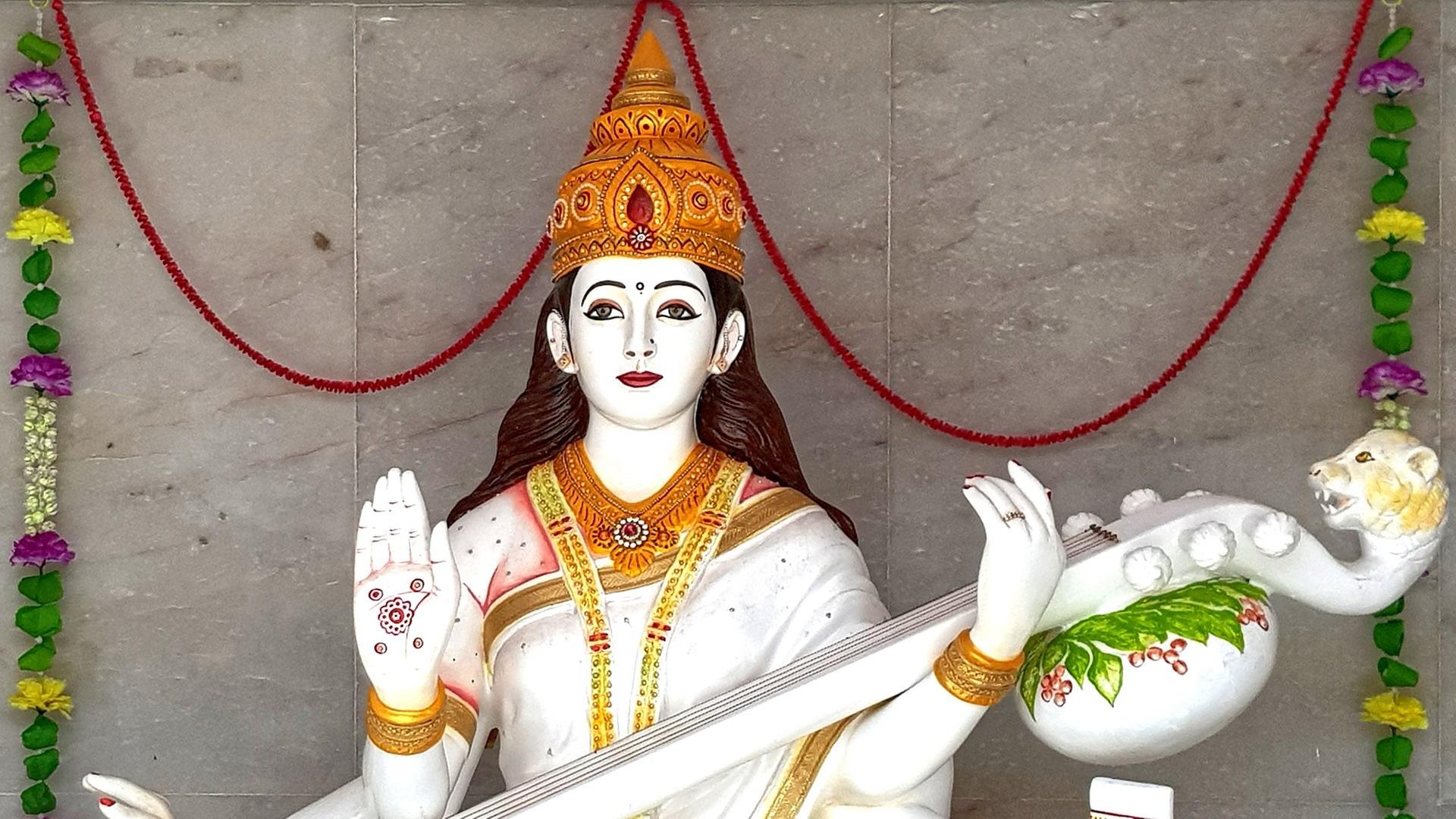 Skulptur der hinduistischen Gottheit Sarasvati, Göttin der Musik, mit goldener Krone und erhobener rechter Hand, in der anderen Hand hält sie ein Saiteninstrument mit Löwenkopf.