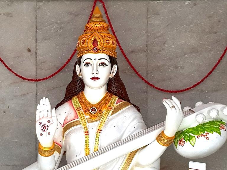 Skulptur der hinduistischen Gottheit Sarasvati, Göttin der Musik, mit goldener Krone und erhobener rechter Hand, in der anderen Hand hält sie ein Saiteninstrument mit Löwenkopf.