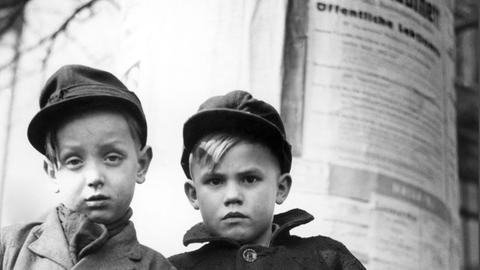 Zwei Berliner Jungs stehen mit skeptischem Gesichtsausdruck und den Händen in den Taschen vor einer Lifaßsäule. Aufnahme aus den 50er-Jahren.