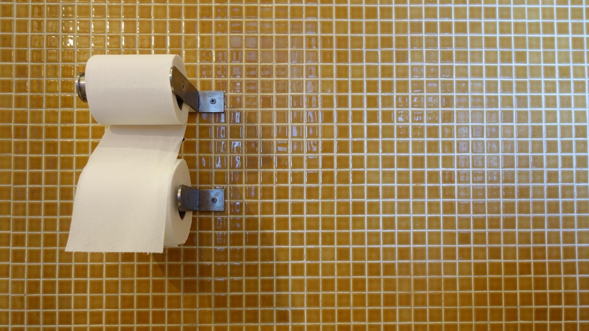 An einem Papierrollenhalter hängen zwei Rollen Toilettenpapier vor einer senfgelb gekachelten Wand. An einem Papierrollenhalter hängen zwei Rollen Toilettenpapier vor einer senfgelb gekachelten Wand.