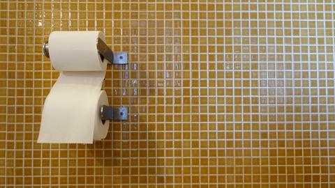 An einem Papierrollenhalter hängen zwei Rollen Toilettenpapier vor einer senfgelb gekachelten Wand.