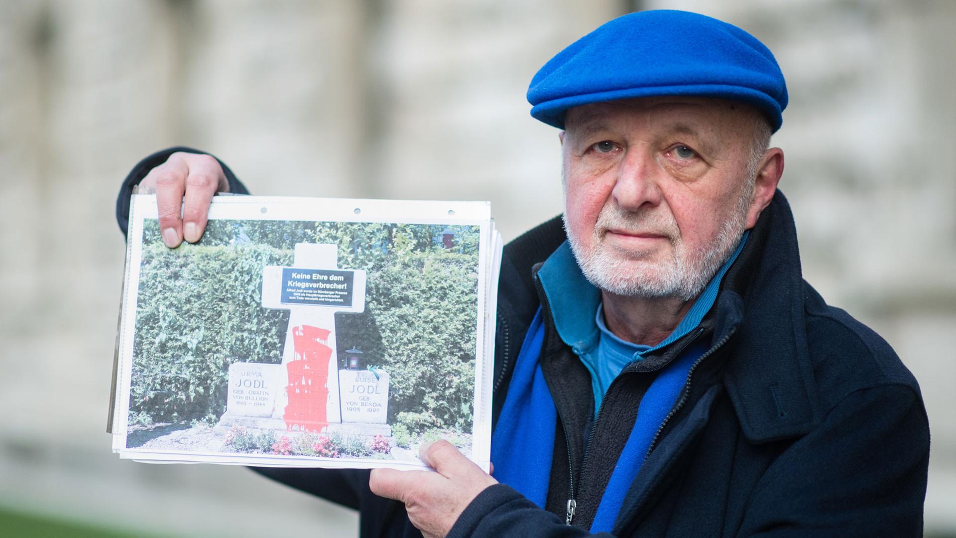 Ein Mann mit blauer Mütze und blauem Schal, der Künstler Wolfram Kastner, hält die Fotografie eines Denkmals, welches mit roter Farbe beschmiert ist, in die Höhe.