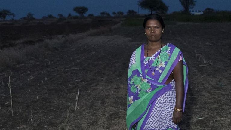 Eine ernst blickende Frau steht in türkis-lilanem Sari bei Nacht auf einem fast kahlen Feld. Eine ernst blickende Frau steht in türkis-lilanem Sari bei Nacht auf einem fast kahlen Feld.