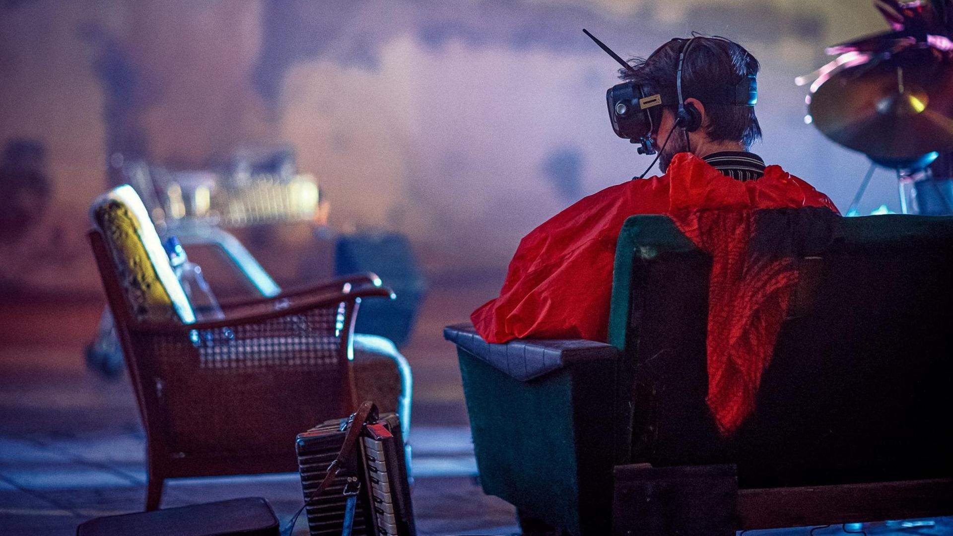 Ein Mann ist von hinten zu sehen, wie er auf einen Sessel sitzt, eine VR-Brille samt Antenne auf dem Kopf trägt und ein auffallend rotes Oberteil. Neben ihm auf dem Boden steht ein Akkordeon.
