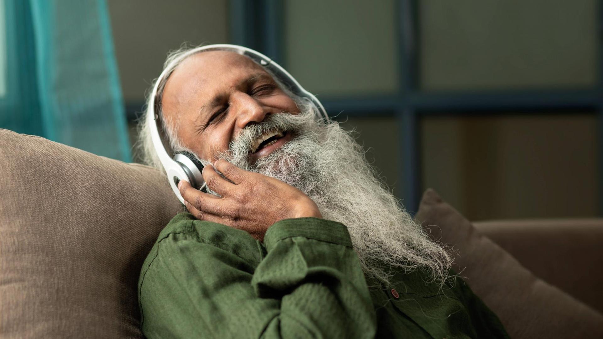 Ein Mann mit weißem Bart hört lachend und glücklich etwas über Kopfhörer.