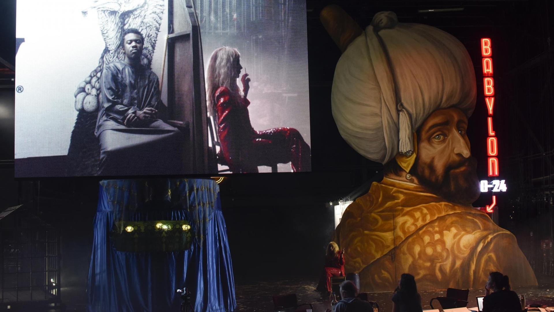 Auf einer Theaterbühne sind ein blaues Zelt und mehrere Videoleinwände mit Aufnahmen sowie eine Schauspielerin zu sehen.