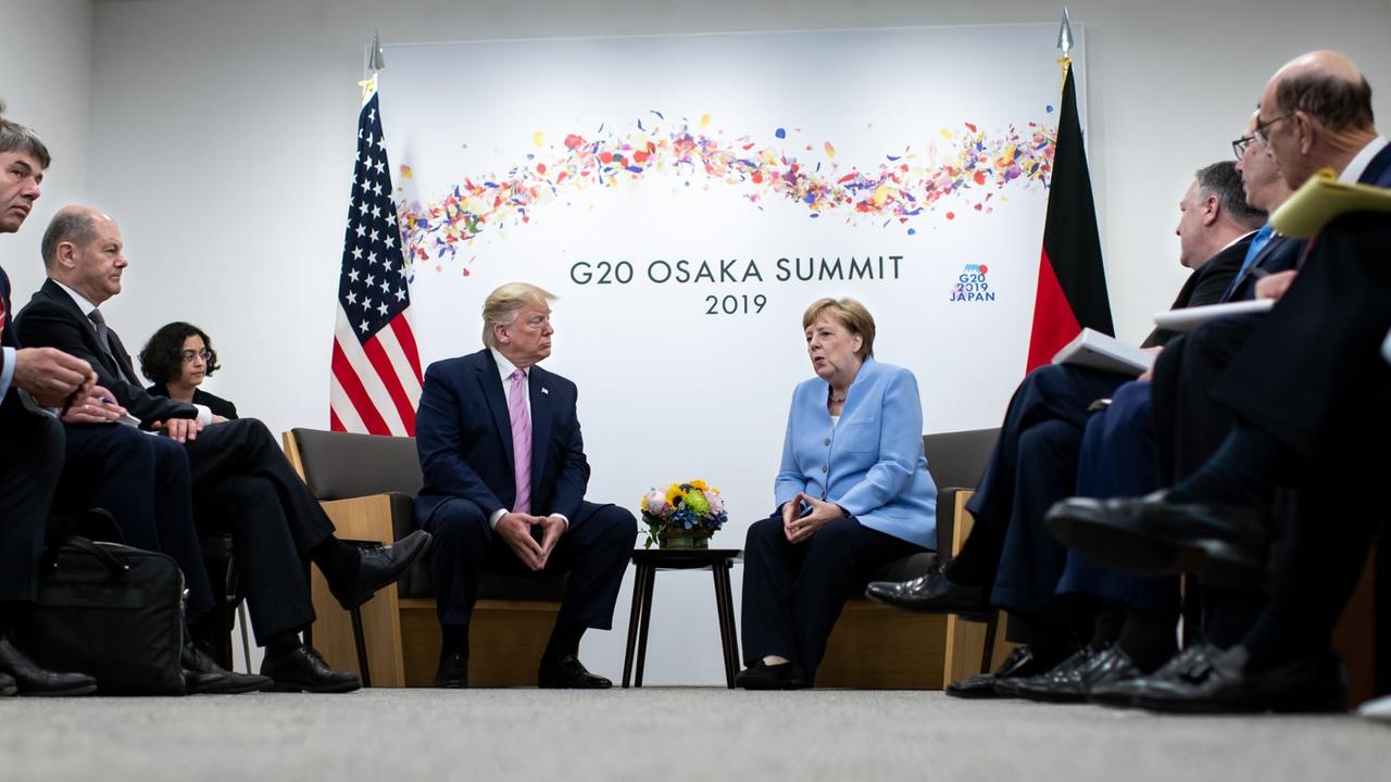 Das Bild zeigt Angela Merkel und Donald Trump. Sie sitzen beide an einem Tisch. Im Hintergrund sind die deutsche und die US-Fahne sowie der Schriftzug des G20-Gipfels in Osaka zu sehen.