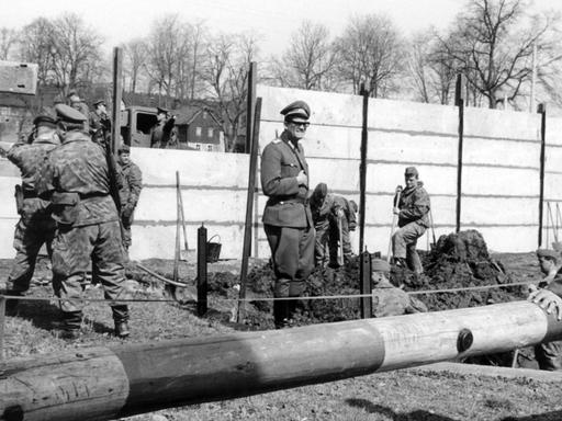Pioniere der Volksarmee der DDR beim Bau der Mauer durch die Ortschaft Mödlareuth. Rechts hinter dem Schlagbaum beobachtet ein Angehöriger des westdeutschen Grenzschutzes die Bauarbeiten an der innerdeutschen Grenze, die mitten durch den Ort führt.