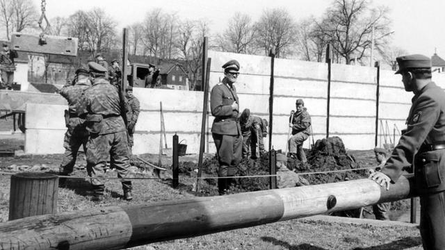 Pioniere der Volksarmee der DDR beim Bau der Mauer durch die Ortschaft Mödlareuth. Rechts hinter dem Schlagbaum beobachtet ein Angehöriger des westdeutschen Grenzschutzes die Bauarbeiten an der innerdeutschen Grenze, die mitten durch den Ort führt.