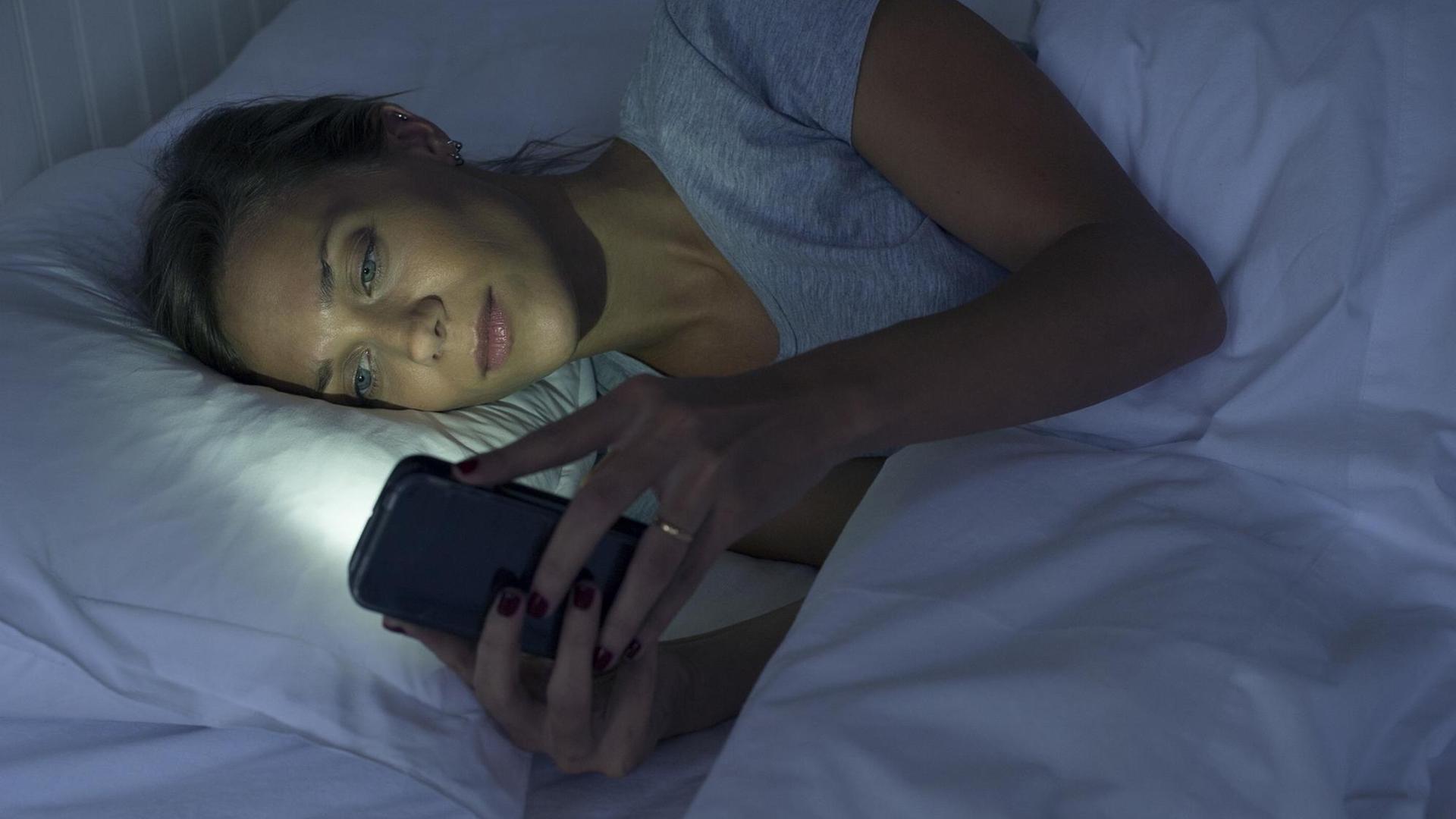 Eine Frau liegt im Dunkeln im Bett und schaut auf das leuchtende Display ihres Smartphones Eine Frau liegt im Dunkeln im Bett und schaut auf das leuchtende Display ihres Smartphones