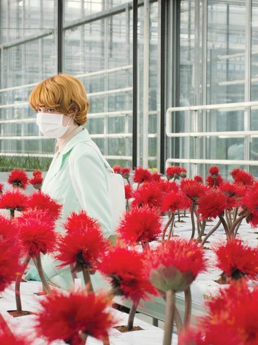 Die Genetikerin Alice (Emily Beecham) mit Mundschutz im Labor, umgeben von roten Blumen.