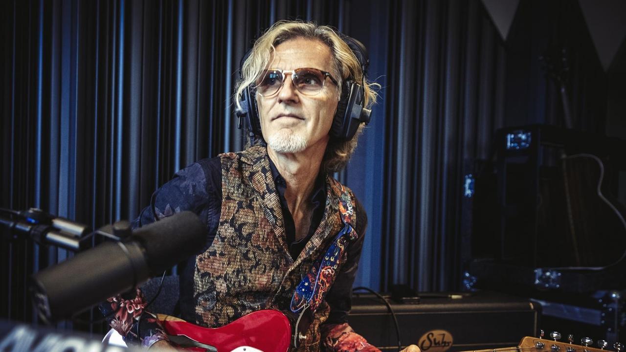 Ein Mann mit blonden Haaren und Brille trägt eine gemusterte Weste. Erhat Kopfhörer auf und hält eine Gitarre in der Hand. Ein Mann mit blonden Haaren und Brille trägt eine gemusterte Weste. Erhat Kopfhörer auf und hält eine Gitarre in der Hand.