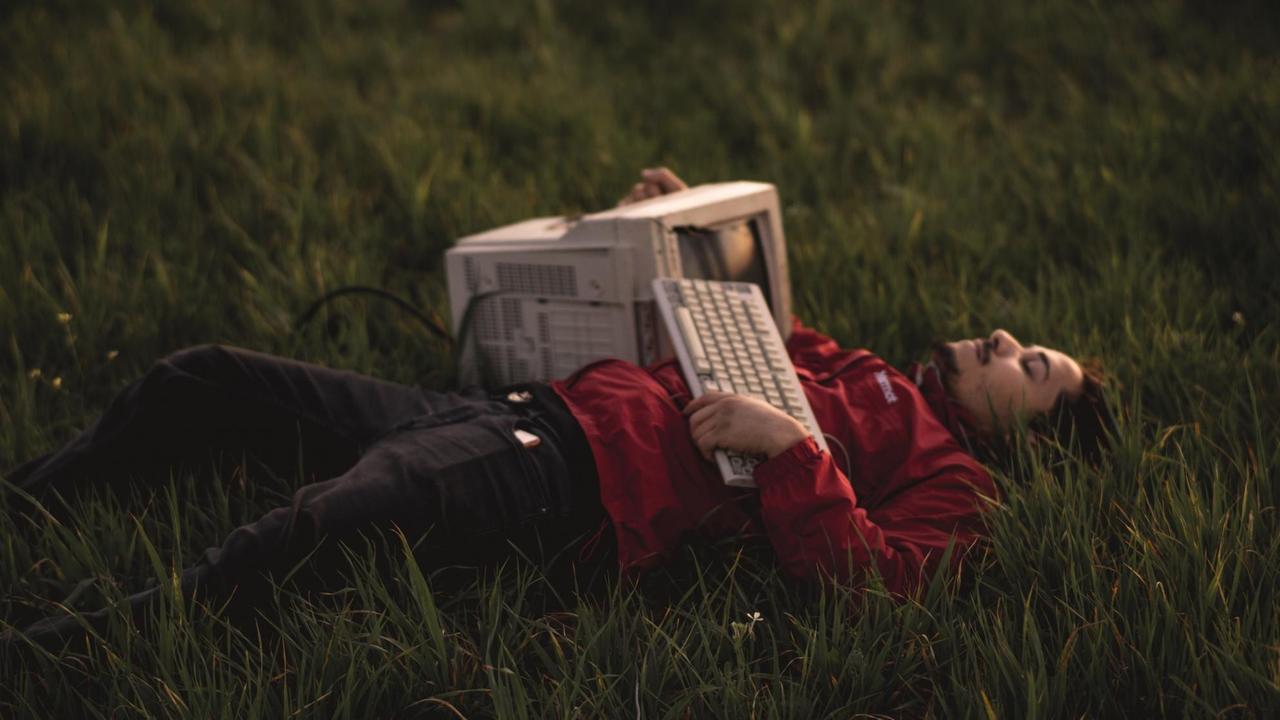 Ein junger Mann liegt mit seinem alten Computer und Tastatur gemeinsam im Gras.