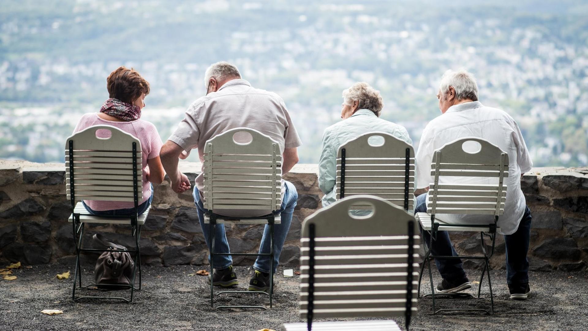 Auf einer Aussichtsplattform stehen vier Stühle auf denen vier ältere Menschen sitzen. Im Hintergrund, in der Unschärfe schaut man auf eine grüne Stadt hinab. Auf einer Aussichtsplattform stehen vier Stühle auf denen vier ältere Menschen sitzen. Im Hintergrund, in der Unschärfe schaut man auf eine grüne Stadt hinab.